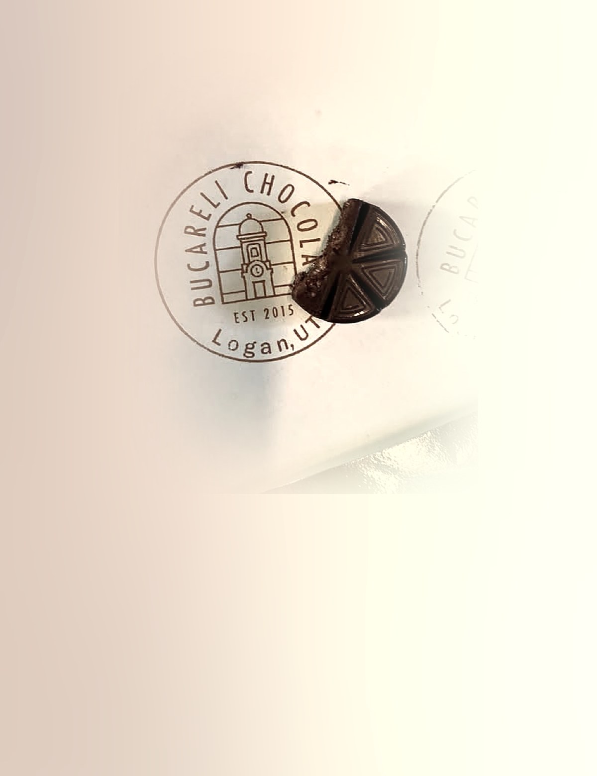 A round Bucareli chocolate tablet, scored into segments, resting on paper printed with the circular Bucareli Chocolate stamp from Logan, Utah.